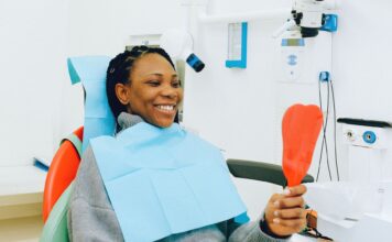 Korony porcelanowe czy licówki kompozytowe – co wybrać, aby poprawić estetykę uśmiechu Uśmiechnięta pacjentka z lusterkiem podczas kontroli u dentysty